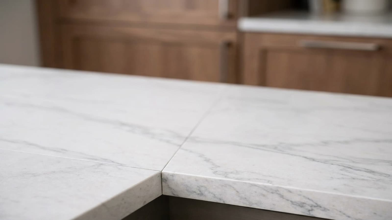 A close view of a seam in a white marble-veined countertop: two pieces of stone meeting in a fine straight joint that runs across the polished surface and continues down through the front edge